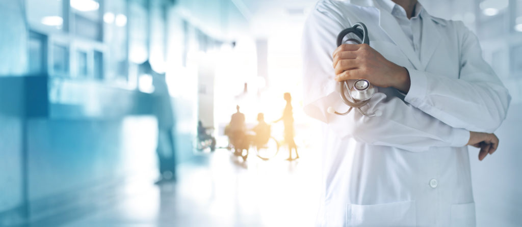 Neck-down photo of a doctor wearing a white coat, crossing his arms and holding a stethoscope in his left hand, symbolizing medical malpractice attributed to Covid-19. There are patients and staff blurred in the background.