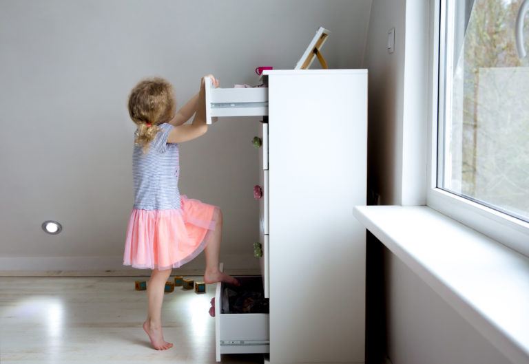 Large photograph of a female toddler girl climbing on a white dresser not affixed to the wall, symbolizing the importance of securing furniture, safety recalls, and tip-over lawsuits.