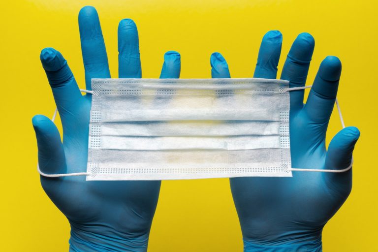 Closeup of a hands wearing bright blue medical gloves, stretching out a white surgical mask, against a bright yellow background, meant to symbolize the impact of COVID-19 on personal injury cases.