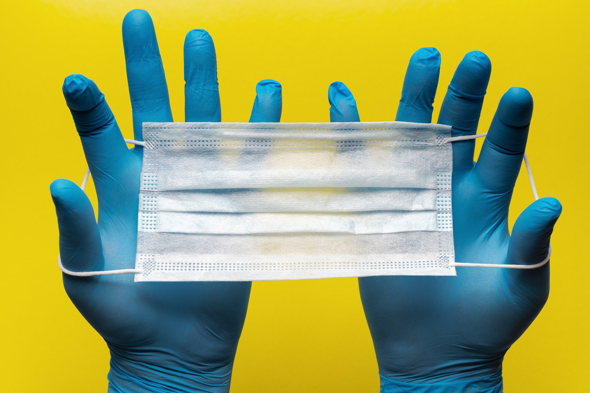 Closeup of a hands wearing bright blue medical gloves, stretching out a white surgical mask, against a bright yellow background, meant to symbolize the impact of COVID-19 on personal injury cases.