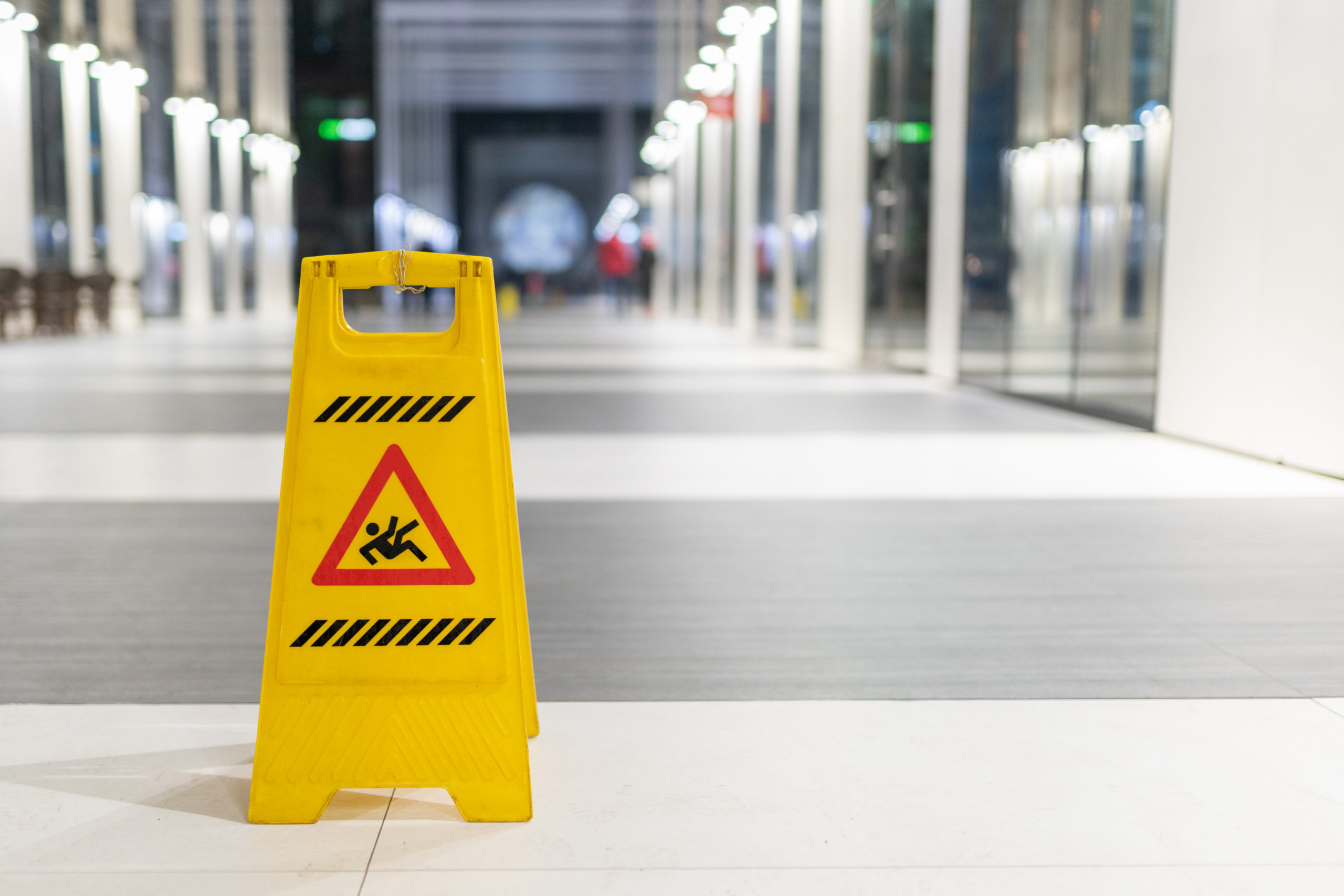 A yellow caution wet floor folding sign in the forefront and on the left, in front of a long broad walkway of smoot grey and white flooring, posing the question, What to do after a slip and fall accident in Philadelphia.
