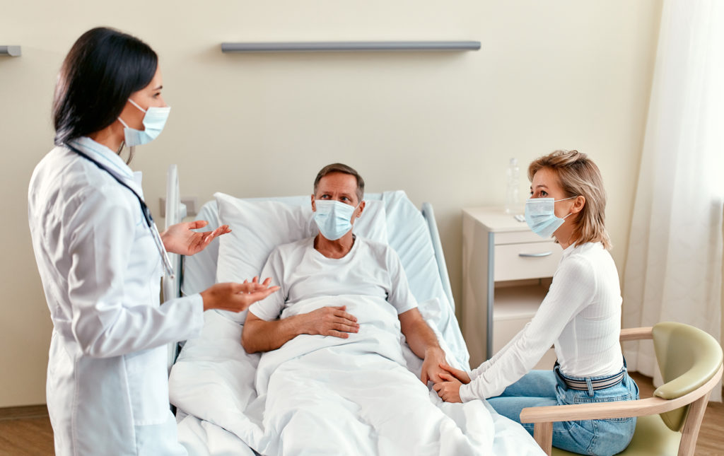 A man recovering from a personal injury in Philadelphia laying in a hospital bed wearing a mask and holding his wife's hand, who is also wearing a mask. A female doctor stands nearby wearing a mask and answerin gthe man's question, Will COVID-19 impact your personal injury case in Philadelphia?