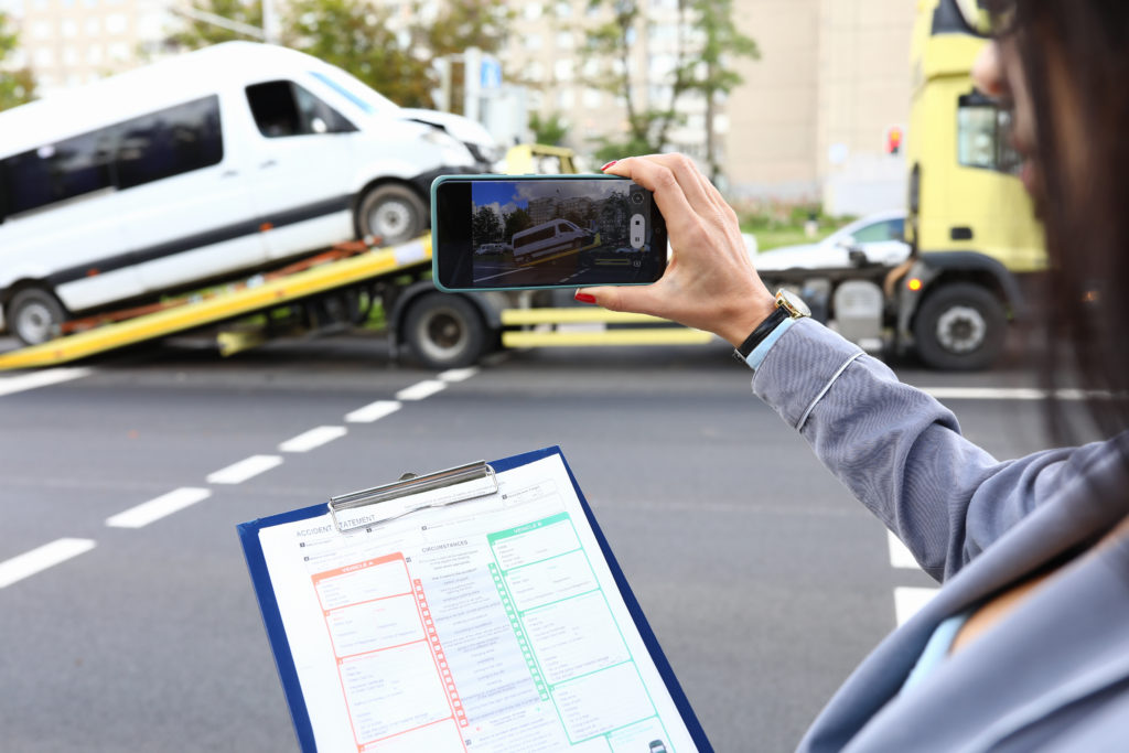 A woman's hand holds a phone out from the right side of the image, to take a picture of a white truck being loaded on to a tow truck in the distance. In the foreground a clipboard with a checklist can be seen, posing the question, will your truck accident case settle in Philadelphia?