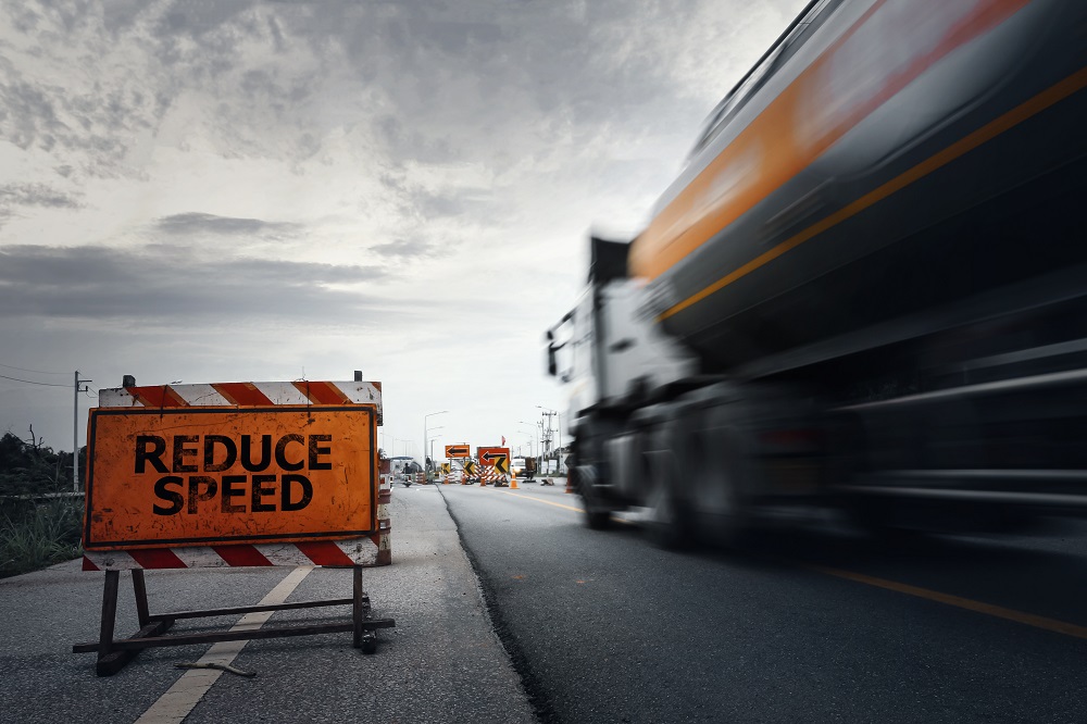 A blur of a semi-truck moves forward on the right, while an orange REDUCE SPEED sign sits on the asphalt road on the left, posing the question, Will your truck accident case settle in Philadelphia?