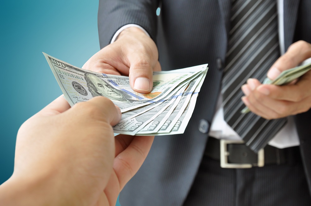 A man in a dark suit, white shirt, and striped grey tie hands money to a pair of receiving hands, posing the question, What is the average payout for a personal injury claim?