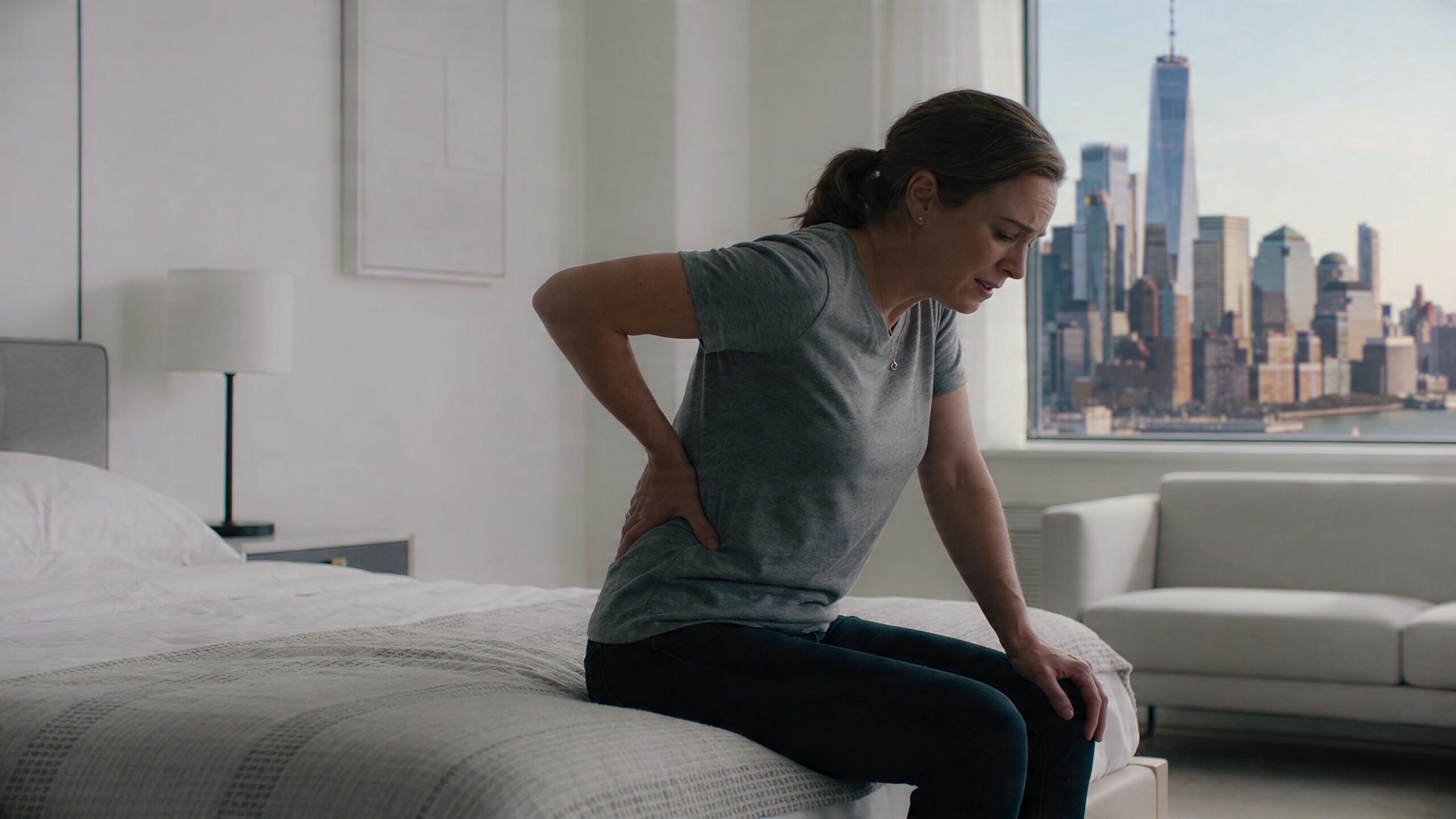 A woman sitting on the edge of her bed feeling pain and discomfort in her lower back.