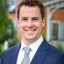 Professional photograph of personal injury attorney John Mattiacci, a young caucasian man with short brown hair, crossing his arms and smiling, wearing a steel-blue suit, white shirt, silver tie, and wedding ring. There is a brick building and green shrubbery in the background.